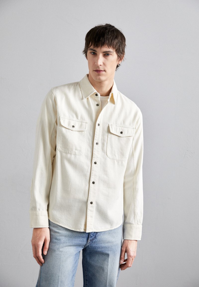 Camisa de botones beige con dos bolsillos en el pecho, botones negros y mangas largas; combinada con jeans azul claro. Textura de tela suave.