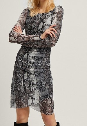 Kobieta ubrana w sukienkę z długim rękawem sięgającą kolan, z czarno-szarym wzorem w skórę węża, z założonymi rękami, na jednolitym tle.