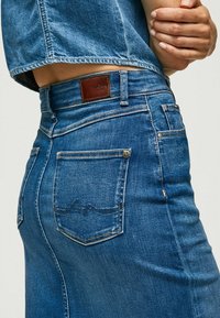 Närbild på en person som bär blå denimjeans och en matchande denimjacka, med händerna korsade vid midjan.