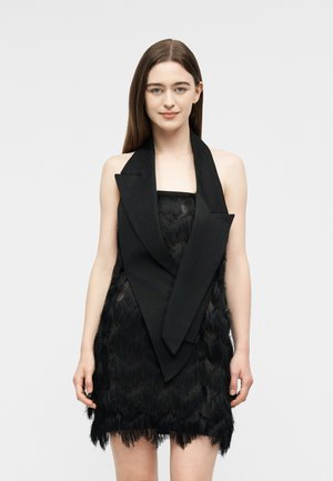 Ženska s ravnimi rjavimi lasmi nosi črno mini obleko brez rokavov z veliko ovratno ovratnico, stoji pred enostavnim svetlim ozadjem.