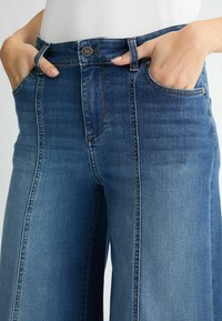 Suurte sääre otste ja kõrge vöökohaga sinised denim püksid, millel on viis taskut ja kontrastsed õmblused, ning mis on varustatud tõmbluku ja nupuga kinnitusega.