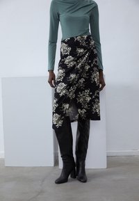 Jupe noire à motifs floraux avec un design enveloppant, assortie à un haut teal à manches longues et des bottes noires à talons montant jusqu'aux genoux. Tissu lisse, motifs élégants.