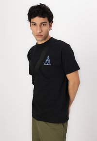T-shirt en coton noir à manches courtes, avec un logo triangulaire brodé bleu sur la poitrine gauche. Le modèle porte une sangle ajustable.