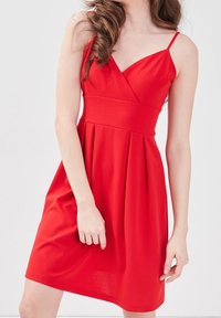 Robe rouge sans manches avec un décolleté en V, taille cintrée et jupe plissée, fabriquée en tissu lisse avec une texture douce.