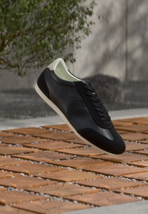 Zapatilla de cuero negro y blanco con cordones flotando sobre una superficie exterior de madera y grava, con un árbol y una pared desenfocados en el fondo.