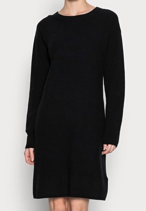 Kobieta ubrana w czarną, prążkowaną sukienkę dzianinową do kolan z długimi rękawami i okrągłym dekoltem na jednolitym jasnym tle.