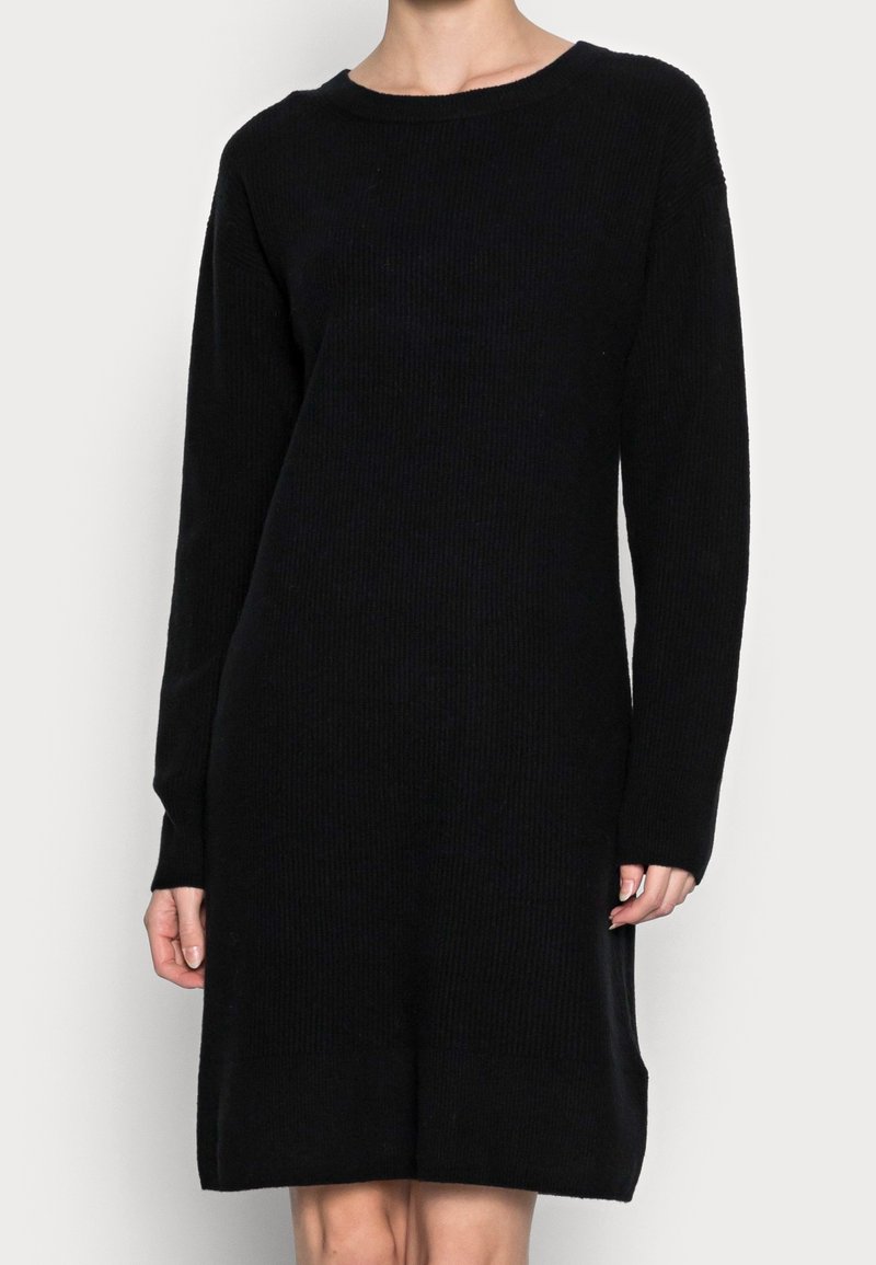 Femme portant une robe noire côtelée en maille, longueur genou, à manches longues et col rond, sur un fond clair uni.