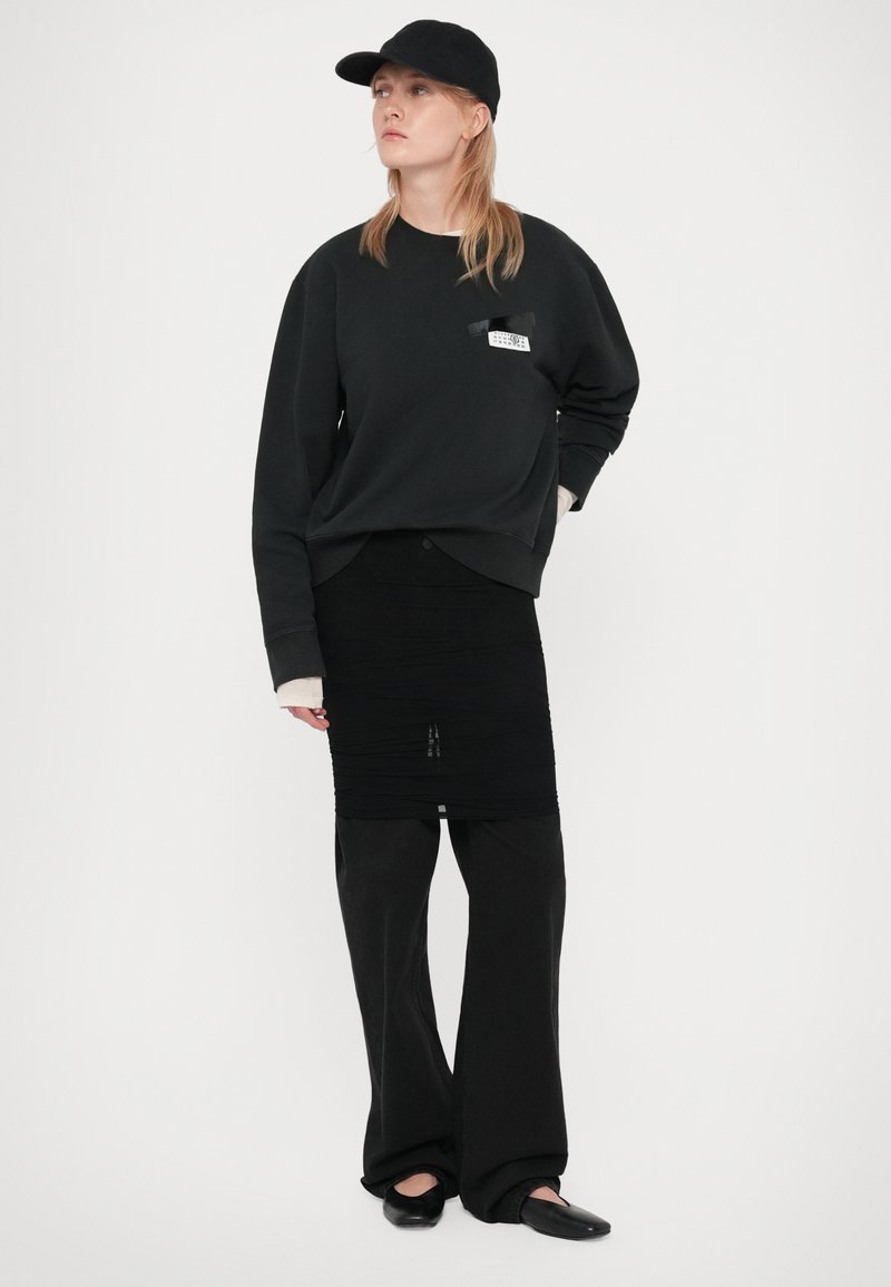 Eine Frau trägt eine schwarze Mütze, einen übergroßen schwarzen Sweatshirt, einen schwarzen gerafften Rock über einer weiten schwarzen Hose und schwarze flache Schuhe, während sie vor einem weißen Hintergrund steht.