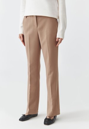 Pantaloni beige su misura con design a gamba dritta, tessuto texturizzato, piega frontale e tasche laterali, abbinati a ballerine nere.