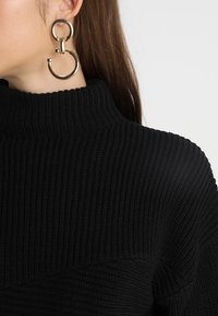 Czarny prążkowany sweter z golfem o teksturowanej tkaninie. Złote kolczyki w kształcie obręczy z unikalnym zapięciem. Włosy ułożone na jedną stronę.