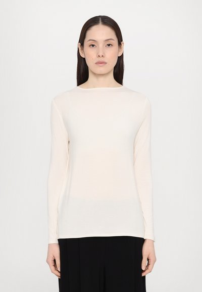 Chemise à manches longues couleur crème en tissu léger et doux, avec un col rond et des fentes sur les côtés, associée à un pantalon noir.
