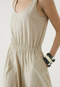 Robe beige sans manches avec une taille élastique. Tissu texturé, tombant à mi-cuisse. Portée avec des bracelets empilés noirs et crème.