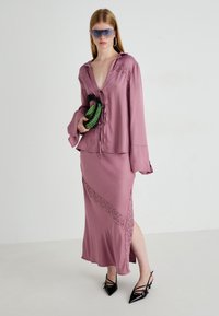 Abito lungo di raso mauve con dettagli in pizzo, abbinato a tacchi neri a punta e una pochette verde texturizzata. La modella indossa occhiali da sole oversize.