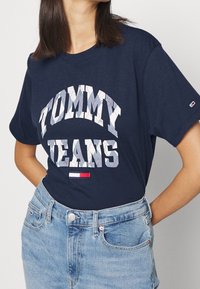 Námořnická modrá bavlněná tričko s výraznou bílou a šedou grafikou "TOMMY JEANS". Obsahuje pruhovanou logo nášivku na lemu, volnější střih a krátké rukávy.