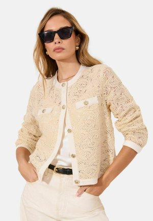 Woman wearing cream lace cardigan with gold buttons, white shirt, light pants, black belt, gold hoop earrings, and black cat-eye sunglasses.