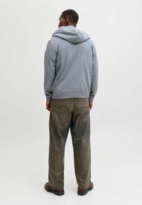 Sweat à capuche en coton gris avec une coupe décontractée et une grande capuche, associé à un pantalon marron délavé et ample ainsi qu'à des chaussures foncées. Design simple, sans motifs.