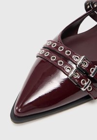 Chaussure en cuir verni bordeaux à bout pointu avec double lanières dotées d'œillets circulaires en argent et de boucles réglables. Texture lisse et brillante.