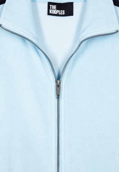Lichtblauwe zip-up jas met een gladde textuur, voorzien van een metalen rits en een zwart label met witte tekst op de kraag.