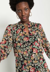 Vestido com estampa floral, mangas longas, base preta e cores incluindo vermelho, amarelo e verde. Detalhe de decote em forma de olho-chave, tecido leve.