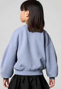 Sudadera oversized de color azul claro con puños y dobladillo acanalados, con una textura suave. Usada con una falda negra, vista desde la parte posterior.