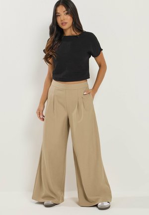 Donna in piedi che indossa un crop top nero a maniche corte, pantaloni beige a gamba larga e vita alta, e sneakers chiari su sfondo semplice.