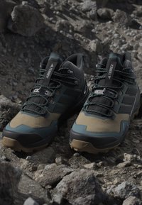 Ciemnozielone i beżowe buty outdoorowe z teksturowanymi powierzchniami, funkcjami stabilizującymi, systemem sznurowania oraz solidną czarną podeszwą na rocky terenie.