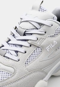White athletic shoe with mesh and suede texture, featuring white laces, Fila branding, and a distinct layered design.