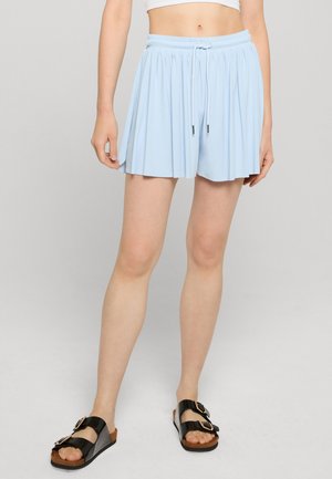 Žena nosi svetlomodré plisované šortky so sťahovacou šnúrkou v páse a čierne sandále so sponou, zobrazená od pása po chodidlá na sivom pozadí.