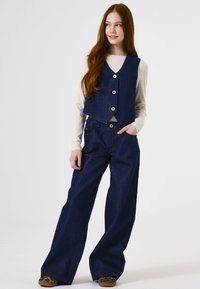 Marineblauer Denim-Westen mit drei Knöpfen, kombiniert mit weit geschnittenen marineblauen Denim-Hosen. Das Model trägt ein hellcremefarbenes Langarmoberteil darunter.