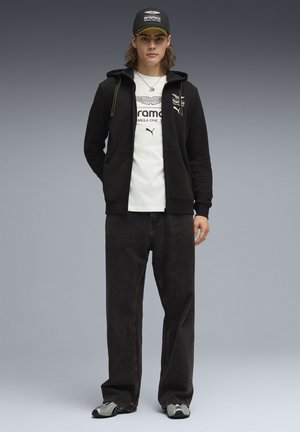 Zwarte capuchontrui met ritssluiting en groene accenten, wit grafisch T-shirt, donkere spijkerbroek en zwarte sneakers. Accessoires zijn een zwarte pet en een zilveren ketting.