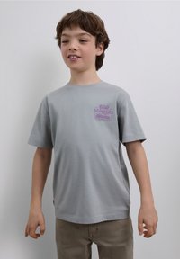 T-shirt en coton gris à manches courtes, avec un texte graphique violet sur le côté gauche de la poitrine. Design simple avec une coupe décontractée.