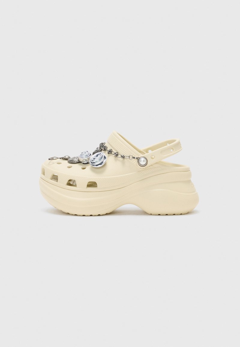 Beige Clogs mit einer klobigen Sohle, die dekorative Charms wie Rosendesigns, eine Perle und Hardware-Akzente an der Vorderseite enthalten.