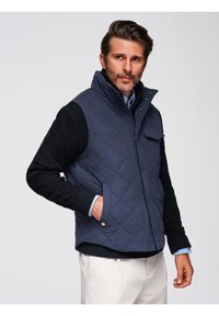 Gilet trapuntato blu navy con colletto alto, hardware dorato e tasche laterali; presenta maniche in maglia nera testurizzata e una camicia azzurro chiaro sotto.