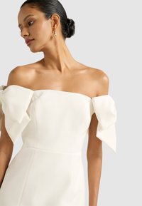 Bijela haljina s jednom naramenicom koja ima velike mašne na rukavima, glatkog materijala i uskog kroja. Jednostavan i elegantan dizajn.