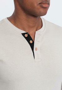 Man som bär en beige henley-tröja med tre träknappar, två på den svarta inre placket, närbild på hals och övre bröst.