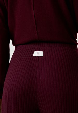 Close-up of a person wearing ribbed, dark burgundy pants and matching sweater with a white label that reads "cool cosy comfy." on the waistband.