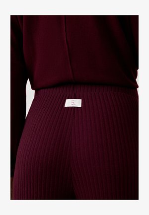 Primo piano di una persona che indossa pantaloni a coste di un profondo borgogna scuro e un maglione coordinato, con un'etichetta bianca che recita "cool cosy comfy" sulla vita.