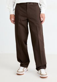 Un pantalon large marron à taille haute, avec une fermeture à bouton sur le devant et une texture lisse, associé à des baskets blanches et beiges.
