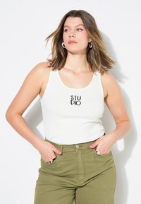 Camiseta sin mangas blanca con franjas y la palabra "STUDIO" impresa en negro, combinada con pantalones de tiro alto color verde oliva, que cuentan con bolsillos delanteros y detalles en plata.