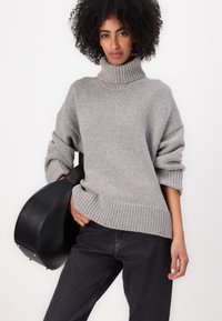 Suéter de cuello alto de punto gris con dobladillo y puños acanalados, combinado con un bolso negro y jeans oscuros. Presenta un ajuste holgado y una textura suave.