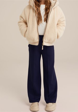 Beige, flauschiger Kapuzenpullover mit Reißverschluss, Kapuze und Taschen vorne, kombiniert mit locker sitzender navyblauer Textilhose und hellbeigen Schuhen.