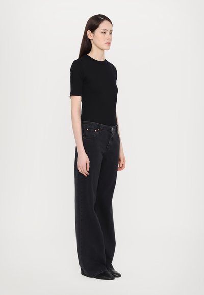 Svart bomullstopp med kort erme kombinert med høyt midje, vide svarte denimjeans. Modellen står foran en nøytral bakgrunn.