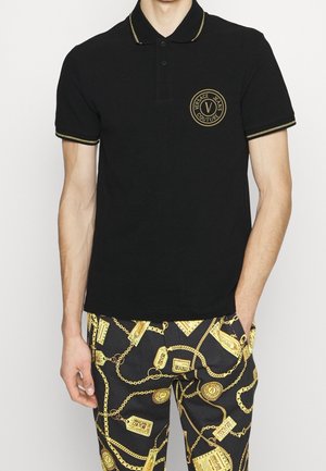 Schwarzes Polohemd mit goldenen Akzenten, versehen mit einem runden Logo auf der linken Brust. Getragen mit gemusterten Hosen, die goldene Ketten und Etiketten zeigen.