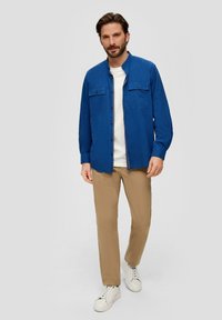 Blauwe button-up shirt met twee borstzakken, gecombineerd met beige broek en witte sneakers. De stof lijkt lichtgewicht en casual te zijn.