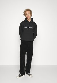 Svart hettegenser med en hvit "carhartt" logo, kombinert med svarte kordfløyelsbukser og svarte joggesko med hvite detaljer.