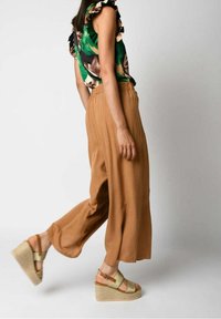 Haut floral vert avec des manches volantées, associé à un pantalon large marron et des sandales à semelle compensée beiges avec une sangle et une texture tissée.