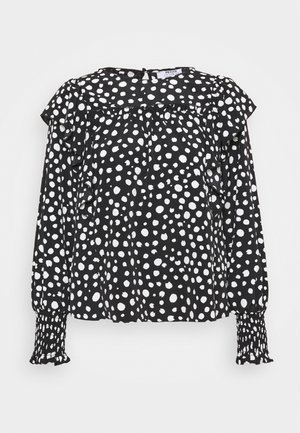 Zwarte blouse met lange mouwen, witte onregelmatige polkadots, ruches over de borst en schouders, en geplooide manchetten.