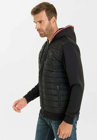 Svart vadderad väst med dragkedja fram, fastsydd svart huva, samt röda och vita detaljer. Har sidfickor och ribbade ärmslut.