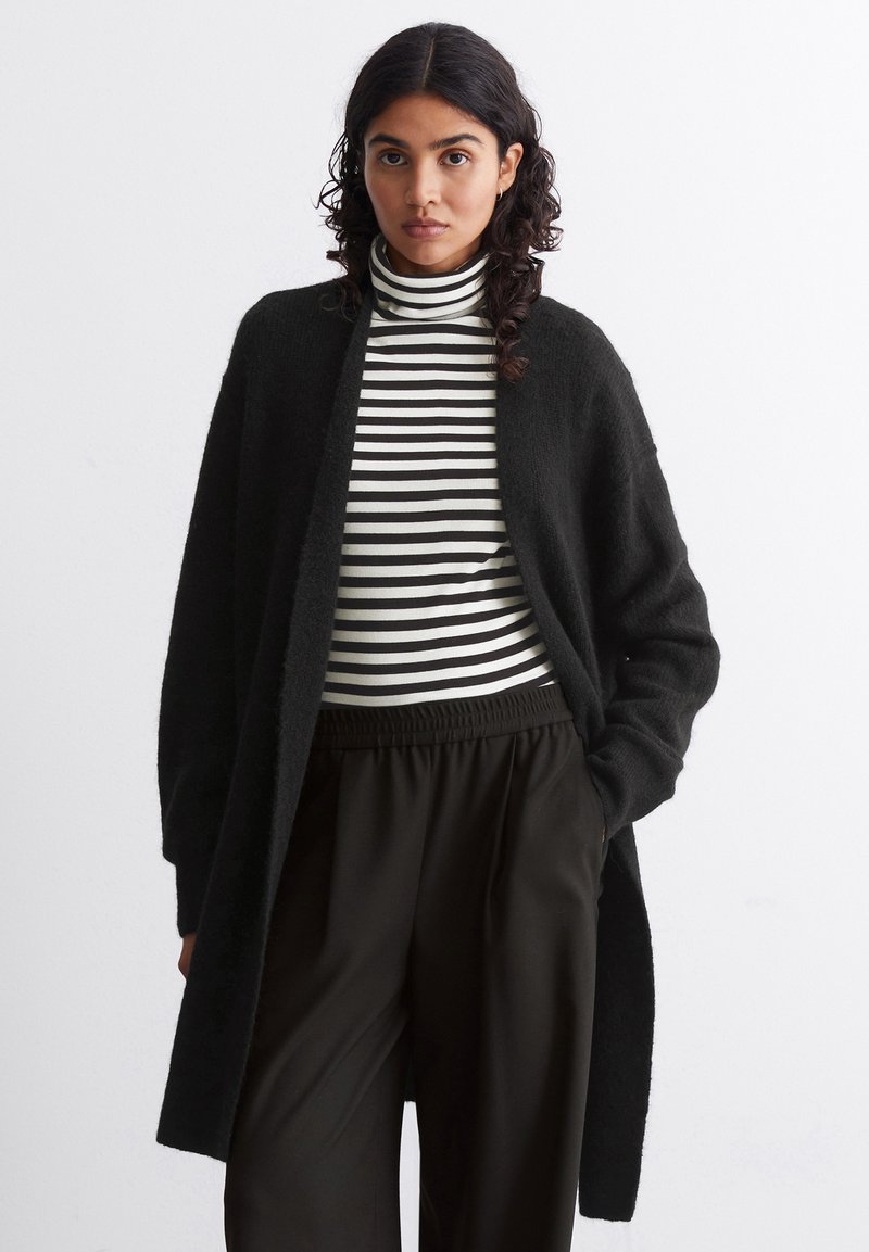 Frau mit lockigem, dunklem Haar, die einen langen schwarzen Cardigan, einen schwarz-weißen gestreiften Rollkragenpullover und lockere schwarze Hose trägt, mit den Händen in den Taschen.