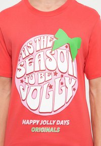 Tricou scurt cu mânecă roșie, prezentând un grafic mare cu textul "Este sezonul să fim veseli" în roz și alb, împodobit cu un accent sub formă de fundă verde.